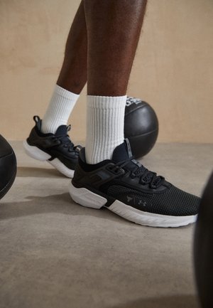 Nogi w czarno-białych butach sportowych i białych skarpetkach stojące na betonowej podłodze obok czarnych piłek lekarskich.