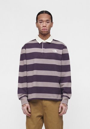 Giovane con vitiligine che indossa una polo a maniche lunghe a righe viola e grigie e pantaloni marroni, in piedi davanti a uno sfondo semplice.