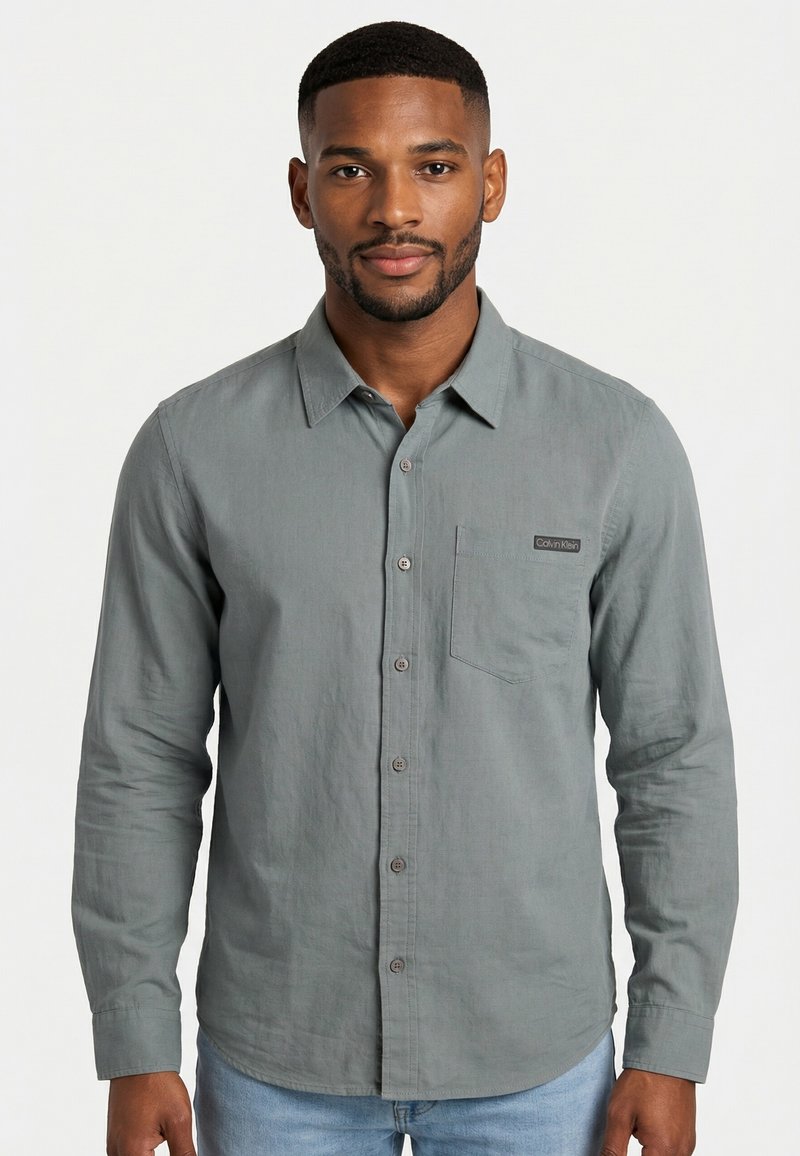 Homme aux cheveux courts et à la barbe, portant une chemise grise à boutons avec une poche poitrine et un jean bleu clair, debout devant un fond uni.