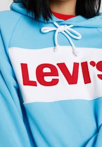 Felpa blu chiaro con fascia bianca che mostra il logo rosso "Levi's". Presenta un cappuccio con cordino e una morbida texture del tessuto.