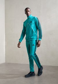 Man in teal tracksuit with black stripes and black sneakers walking indoors against beige wall background.