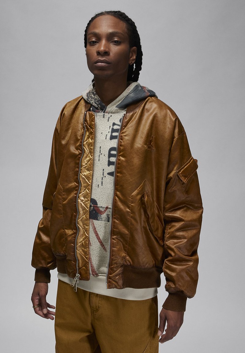 Person mit geflochtenem Haar, die eine braune Lederbomberjacke über einem bedruckten Hoodie und braunen Hosen trägt, vor einem neutralen grauen Hintergrund.