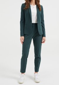 Eine Frau trägt einen dunkelgrünen Maßanzug mit einem weißen T-Shirt und weißen Turnschuhen, stehend vor einem schlichten weißen Hintergrund.