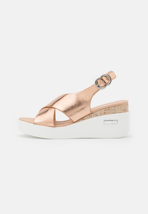 Rose gold metallic wedge sandal with white platform sole, cork detail, cross straps, and silver buckle on heel strap.
