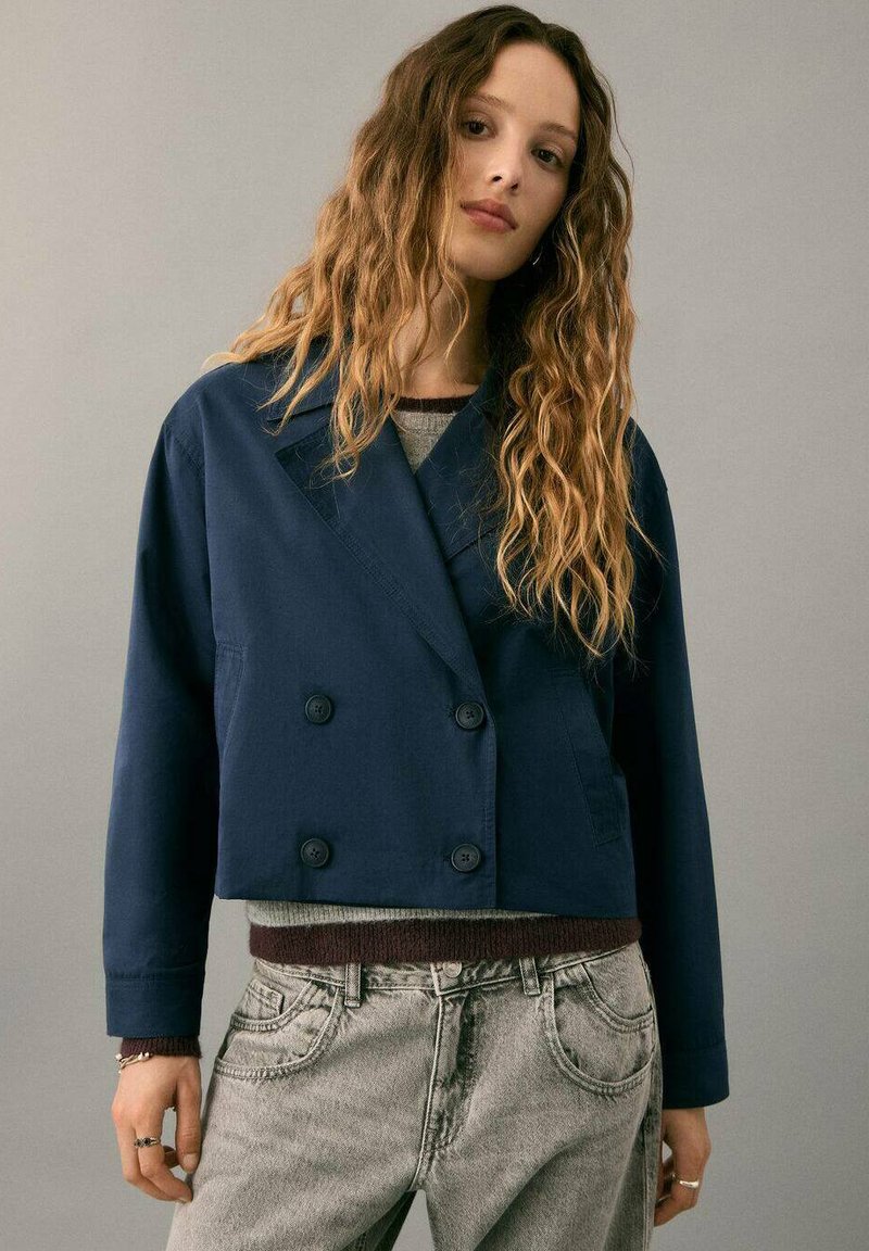 Chaqueta corta azul marino de doble botonadura en una tela suave, con botones y cuello con solapas. Combinada con vaqueros gris claro.