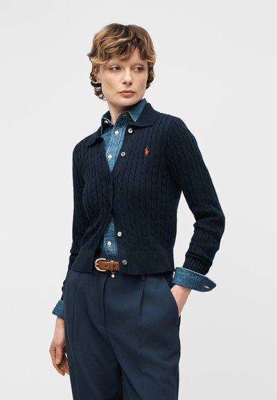 Άτομο που φοράει μπλε ζακέτα με κοτσίδες πάνω από ένα τζιν πουκάμισο, μασχαλισμένη σε μπλε παντελόνια ψηλόμεσα με καφέ ζώνη, με τα χέρια στις τσέπες.