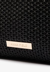 Svart vävt tygväska med en guldtonad metalletikett graverad med "ANNA FIELD." Texturerad yta med en strukturerad form.