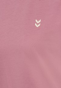 Różowa tkanina z dwoma małymi białymi haftowanymi oznaczeniami w kształcie litery V w pobliżu górnego środka.