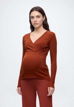Femme enceinte aux longs cheveux foncés porte un haut cache-cœur à manches longues couleur rouille et un pantalon assorti, debout devant un fond blanc uni.