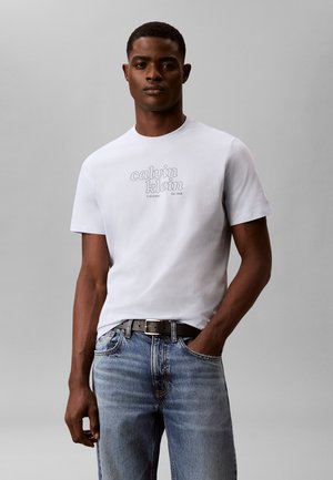 Man draagt een wit Calvin Klein T-shirt en blauwe spijkerbroek, staat met één hand in de zak tegen een eenvoudige grijze achtergrond.