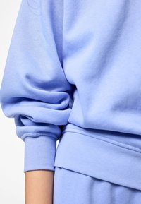 Manche de sweat-shirt bleu clair relevée sur un bras, montrant le poignet et une partie de la ceinture assortie sur un fond blanc.