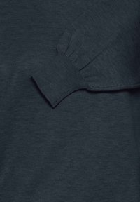 Sötét teal színű kötött pulóver ujja bordás mandzsettával, amely egy azonos színű pulóver test háttérre fekszik.