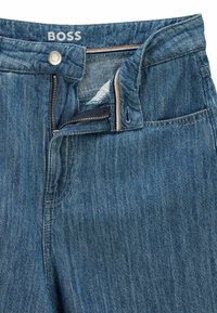 Jeans i denim i en medelblå färg med dragkedja och knappstängning. Har en slät yta och synliga sömdetaljer.