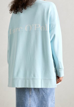 Kobieta o kręconych włosach, ubrana w jasnoniebieską, oversize bluzę z napisem "Marc O’Polo" na plecach oraz jeansową spódnicę.