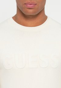 Hombre vistiendo un suéter de punto blanco con el logo de GUESS en relieve, recortado desde la nariz hasta la parte superior del pecho sobre un fondo liso.