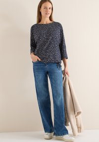 Haut bleu marine avec motif abstrait clair, coupe décontractée et manches trois-quarts, accompagné d'un jean bleu ample et de baskets blanches, tenant une veste beige.