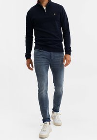 Sudadera navy con cremallera y sección superior texturizada, combinada con jeans ajustados de color azul claro y zapatillas blancas. Acento del logo dorado en la sudadera.