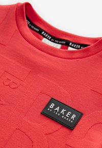 Raudona medvilninė marškinėliai su tekstūruotu logotipu ir juodu stačiakampiu etiketė su balta užrašu „BAKER by TED BAKER.“