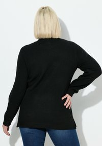 Blonde Frau mit Bobfrisur, die einen schwarzen Langarm-Pullover und blaue Jeans trägt, steht mit einer Hand auf der Hüfte und blickt von der Kamera weg.