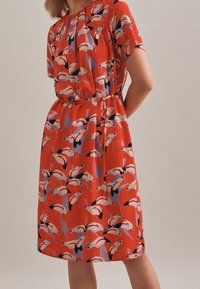 Röd klänning med ett upprepat flamingomönster i nyanser av rosa, svart och lila. Tillverkad av lättviktsmaterial med en avslappnad passform.