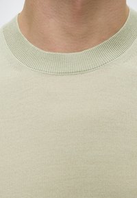 Gros plan sur le cou et le haut de la poitrine d’une personne portant un pull en maille beige clair à col rond.
