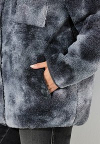 Giacca in fleece nelle tonalità di grigio con una superficie morbida e texturizzata. Presenta una tasca sul petto e tasche laterali, con rifiniture scure lungo i bordi.
