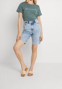 Grön bomullst-shirt med vit text, högmidjade jeansshorts i ljusblått med fransig fåll och beige slip-in-sandaler med vita ränder.