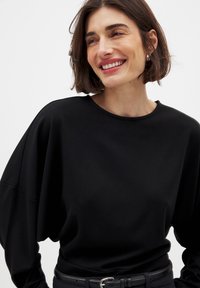 Schwarzer Pullover mit weiten, drapierten Ärmeln und rundem Ausschnitt. Weiches Material mit glatter Struktur. Tragen mit einem schwarzen Gürtel.