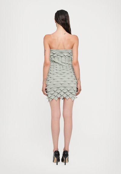 Vestido sin tirantes, ajustado, en verde claro con escamas texturizadas y tridimensionales. El dobladillo tiene un borde irregular. Se lleva con zapatos de tacón alto negros.