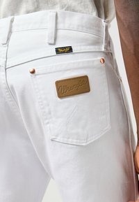 Jean en denim blanc avec des accessoires en cuivre, une écusson en cuir marron, et des accents de couture visibles sur les poches arrière.