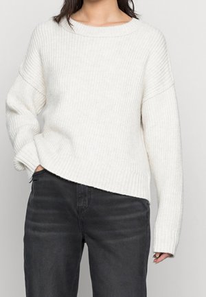 Femme portant un pull tricoté côtelé blanc et un jean noir taille haute, une main dans la poche, sur un fond clair.