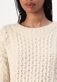 Beige gestrickter Pullover mit einer strukturierten Rippstrickmusterung und einem weiten Rundhalsausschnitt. Der Stoff wirkt weich und leicht flauschig.