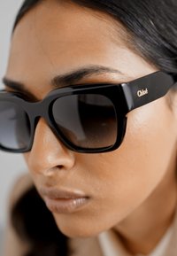 Black oversized sunglasses made of glossy plastic, featuring a square shape and subtle gold branding on the temple. Dark tinted lenses.