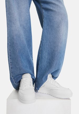 Białe sneakersy z teksturowanej skóry, okrągłym noskiem i płaską gumową podeszwą, zestawione z luźnymi, niebieskimi dżinsami. Buty mają wiązanie na sznurowadła.