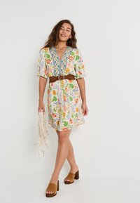 Vestido corto floral en blanco con patrones coloridos, escote en V y bordado azul. Cinturón marrón con hebilla redonda, combinado con zapatos de tacón cuadrado marrones.