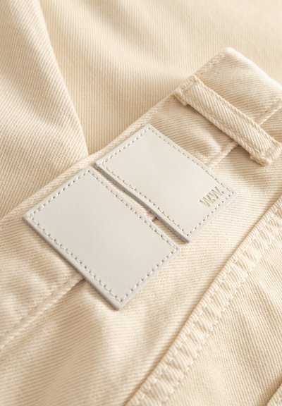 Béžová tkanina s rebrovanou textúrou, na ktorej sú dve obdĺžnikové biele kožené štítky s prešitými okrajmi, reliéfnymi detailmi a bočnou slučkou.