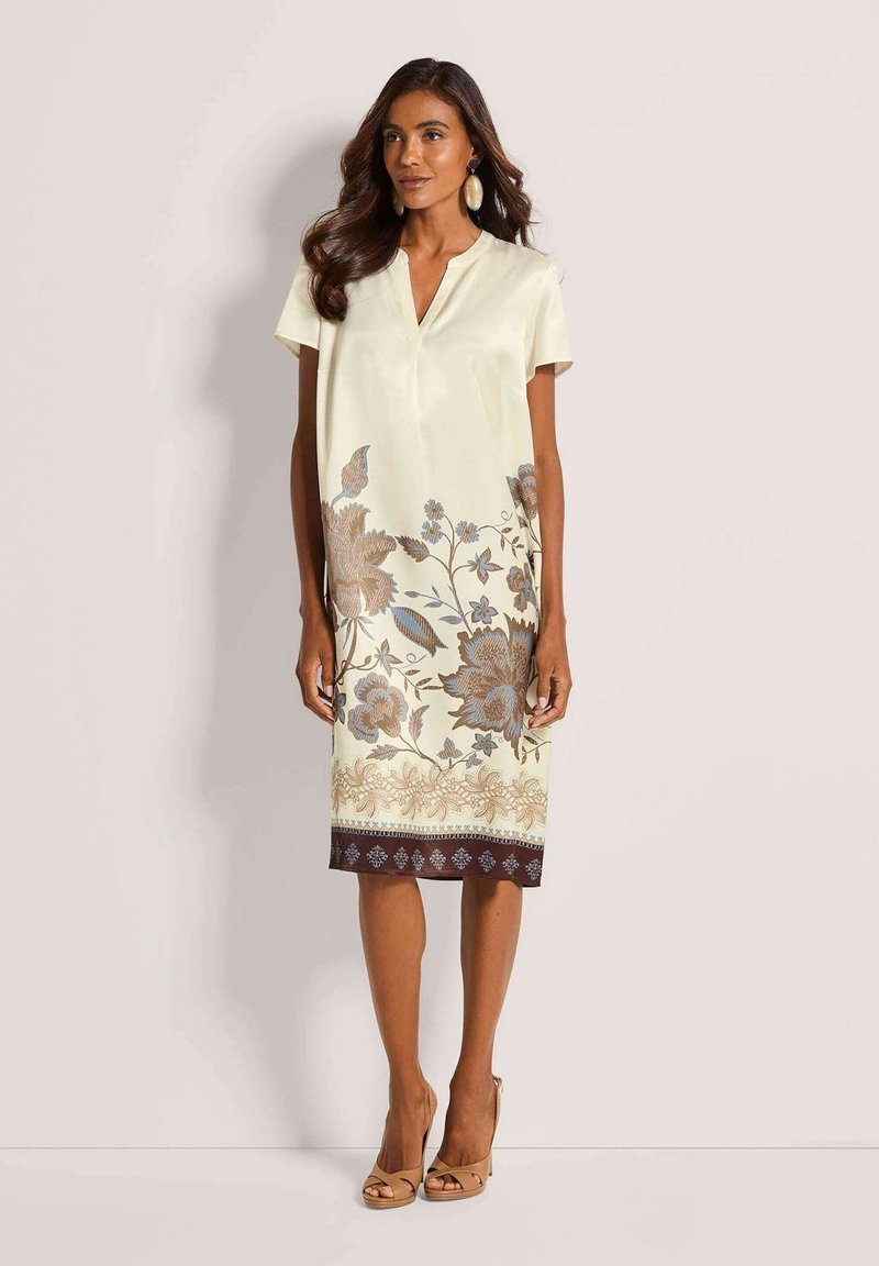 Femme portant une robe crème longueur genoux avec un imprimé floral marron sur la moitié inférieure, manches courtes, col en V, talons beiges et grandes boucles d'oreilles ovales.