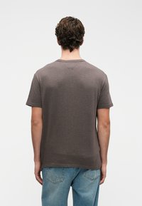 Muž zobrazený zezadu, oblečený v obyčejném tmavě šedém tričku s krátkým rukávem a světlých modrých džínách na b�ílém pozadí.