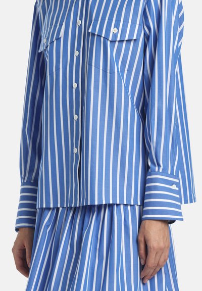 Zils un balts vertikāli strīpainām pogām krekla ar divām krūšu kabatām un atbilstoša strīpainā svārku komplekts.