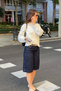 Kootud kreemjas kampsun tekstuuriga disainiga, tume denim culottes ja heledad kralid, hoides telefonit tänavakolmnurgas seistes.