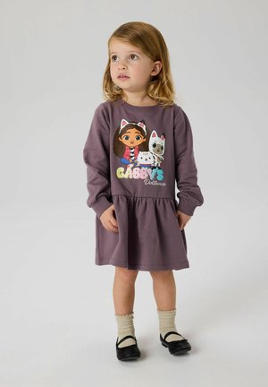 Abito di cotone viola con gonna arricciata, che presenta una grafica colorata di personaggi dei cartoni animati sul petto. Scarpa nera e calzini color crema.