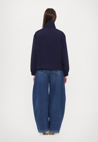 Navy blue jacket with a high collar, elastic cuffs, and a relaxed fit, paired with wide-leg blue denim pants and tan shoes.
