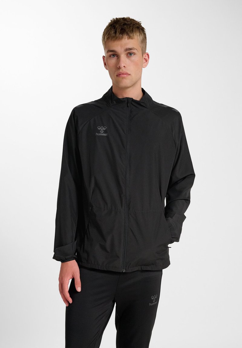 Junger Mann, der eine schwarze Jacke mit Reißverschluss und passende schwarze Hose trägt, steht mit einer Hand in der Tasche vor einem weißen Hintergrund.