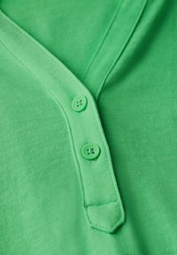 Grön bomullspiké med V-hals, utrustad med tre runda gröna knappar och en ribbad krage för textur.