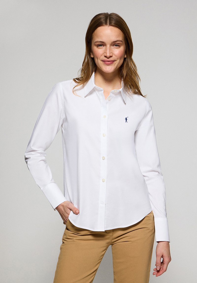 Camicia bianca con bottoni, realizzata in cotone, maniche lunghe, con logo blu navy sul petto, abbinata a pantaloni khaki.