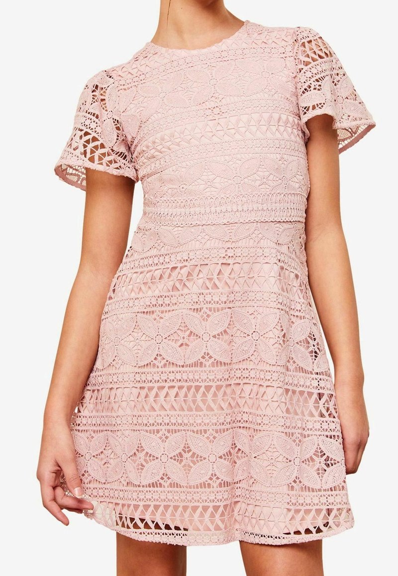 Femme portant une robe en dentelle rose à manches courtes, longueur au genou, avec des motifs floraux et géométriques et un col rond.