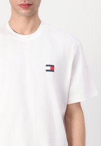 Hvid bomulds t-shirt med korte ærmer, med et kvadratisk logo i marineblå, rød og hvid på venstre brystområde. Rund halsudskæring.