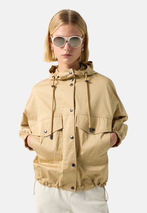 Woman wearing beige hooded jacket with drawstrings and snap buttons, sunglasses, and earrings, standing with hands in pockets.