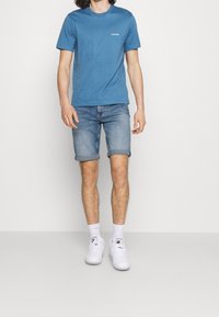 Blue cotton T-shirt with round neck and small white logo, paired with rolled denim shorts and white sneakers. Model standing against a plain background.