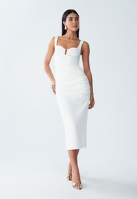 Valkoinen bodycon-mekko sydänmallisella kaula-aukolla, rypytetyillä yksityiskohdilla ja avara etuosa, yhdistettynä kultaisiin korsettikenkäisiin. Suunnittelussa on sileä pinta.